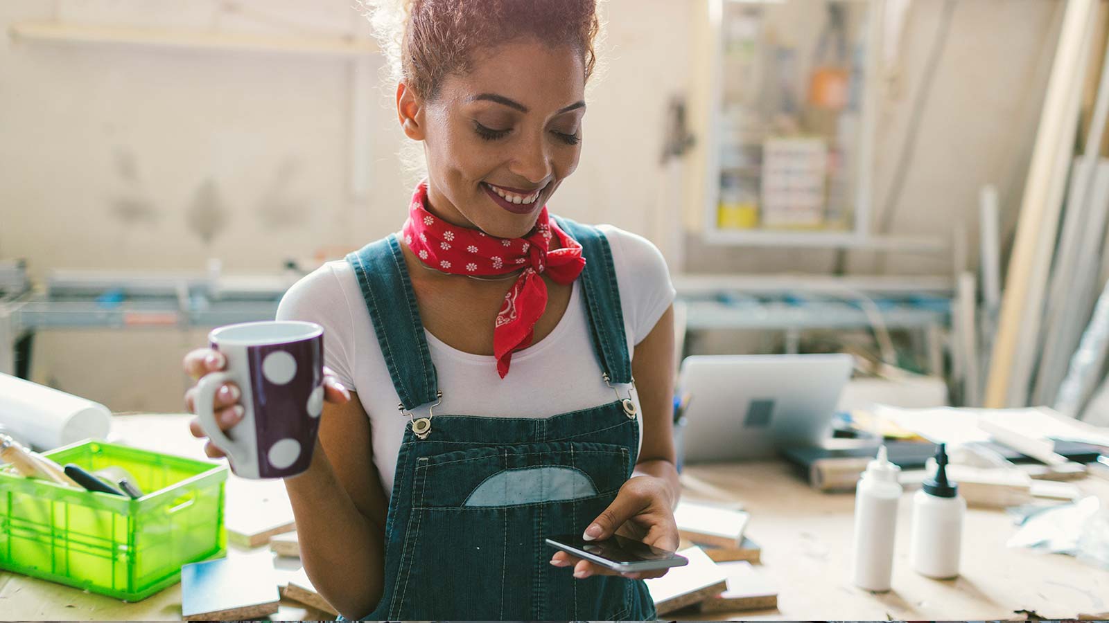 Young woman holding a cup of coffee while looking at her smartphone in her workshop